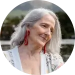 Older individual with long gray hair gently tilts their head, wearing red earrings and a white floral-patterned garment.