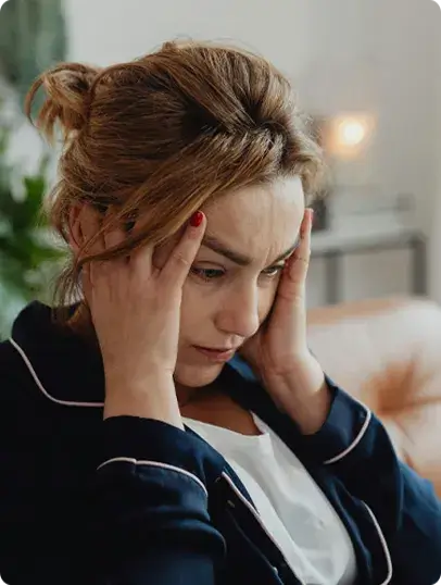 A person sits indoors with both hands pressed to the sides of their head, appearing focused or stressed. They have light brown hair tied back and wear a dark top with white piping over a white shirt. A candle and green plants are softly blurred in the background, suggesting a calm home setting.