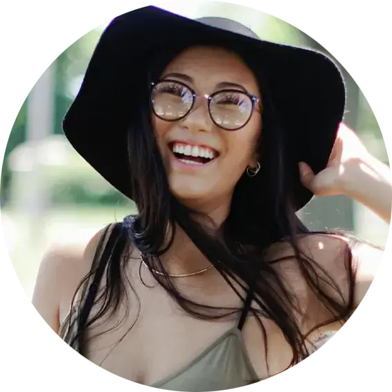 Close-up portrait of a person with long dark hair, wearing a wide-brimmed black hat and large round eyeglasses. They are smiling and looking slightly to the side.