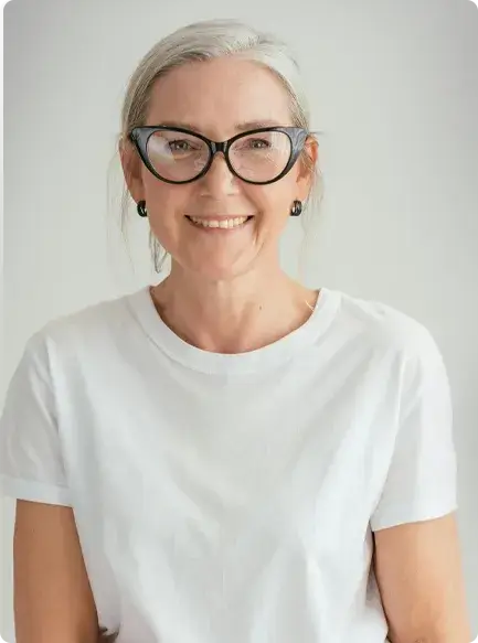Portrait of a person with gray hair, wearing black-framed glasses, small black earrings, and a white t-shirt.