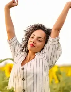 A person with curly hair stands in a field of blooming sunflowers, arms raised and eyes closed. They wear a white shirt with vertical black stripes and buttons down the front. The bright sky and vibrant sunflowers create a serene, joyful atmosphere, suggesting a moment of peace and connection with nature.