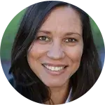 lose-up portrait of a person with long dark hair, smiling and looking directly at the camera. The image is framed in a circle with a softly blurred green-toned background, suggesting an outdoor setting.