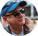 A person wearing a blue cap with yellow text, sunglasses, and a gray jacket smiles as a light-colored dog affectionately licks their cheek.