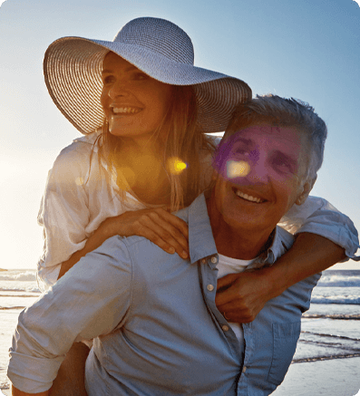 Two people enjoying a joyful moment at the beach during sunset. One person, wearing a wide-brimmed hat, is playfully riding on the back of the other, who is smiling. 