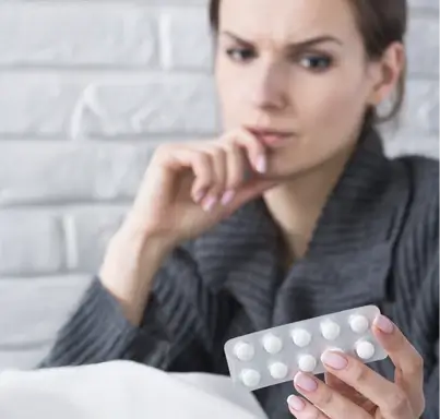 A person wearing a dark gray sweater holds a blister pack of white round tablets, examining them thoughtfully.