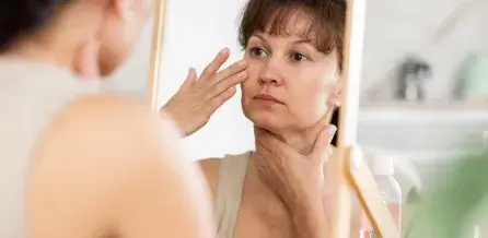 A person closely examines their face in a mirror, touching their cheek with one hand and resting the other under their chin.