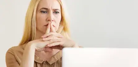 A person with long blonde hair sits in front of a white laptop, hands clasped in front of their face, appearing focused on the screen.