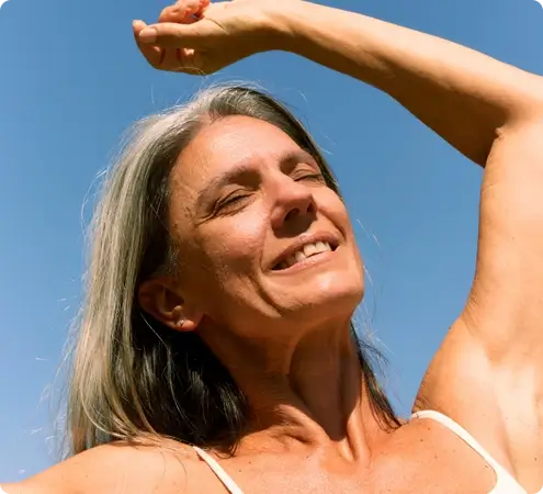 An older individual with long gray hair stands outdoors under a clear blue sky, wearing a light-colored sleeveless top. With one arm raised and eyes closed, they appear relaxed and peaceful, basking in bright sunlight.