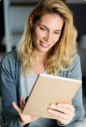 A person with long blonde hair, wearing a gray cardigan over a white shirt, holds and looks at a tablet device. Their expression suggests engagement with the screen, possibly reading, browsing, or video chatting.