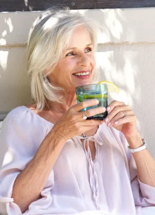 Older individual with light-colored hair sits outdoors against a light wall, smiling and holding a glass of beverage with lemon slices and mint. They wear a light, flowy blouse along with a ring, necklace, and watch.